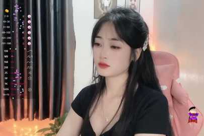 Media: Video of a young Asian woman with long black hair in pigtails, wearing a black top, sitting in a pink chair. Background shows a black curtain and a framed picture.