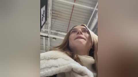 Media: Video of a blonde woman with a slight smile, wearing a white fluffy robe, standing in a warehouse with a corrugated metal ceiling and industrial lights.