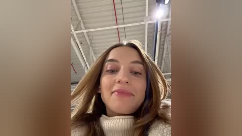 Media: Video of a young woman with light skin, straight brown hair, and fair complexion, wearing a beige turtleneck sweater, standing in a warehouse with exposed metal beams and pipes, looking directly at the camera.