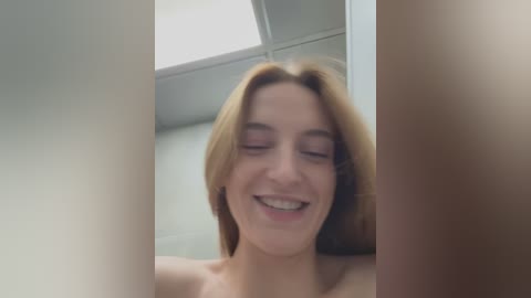 Media: Video of a smiling, topless young woman with light skin and shoulder-length blonde hair, set against a white-tiled bathroom background with a fluorescent ceiling light.