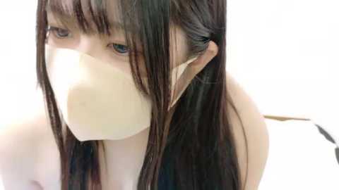 Media: Video of a young Asian woman with long, straight black hair, wearing a white surgical mask, sitting against a plain white background.