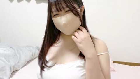 Media: A video of an Asian woman with long dark hair, wearing a white mask and a white off-shoulder top, smiling, in a minimalistic room with white walls and a bed.