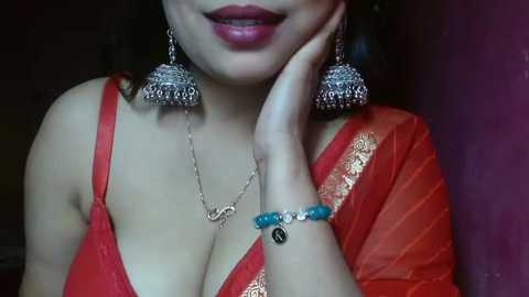 Media: A video of a woman with a light brown complexion, wearing a red saree with gold embroidery, silver jewelry, and a turquoise bracelet, smiling with her hand on her cheek.