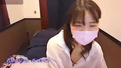 Media: A video shows an Asian woman in a white shirt, with a mask covering her face, lying on a bed with dark sheets. The room has beige walls and a dark curtain.