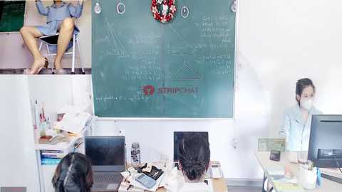 Media: Video of an Asian woman in a blue dress and mask seated at a desk, surrounded by messy office supplies and a computer. Another woman stands by a chalkboard with math formulas, wearing a mask.