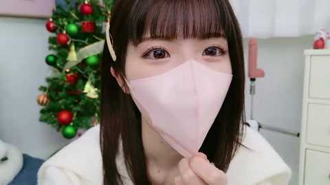 Media: Video of an Asian woman with straight brown hair and bangs, wearing a pink surgical mask, standing in a festive room with a decorated Christmas tree and white furniture.