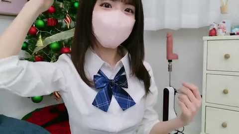 Media: A video of an Asian woman with a white mask, wearing a blue plaid bow tie, in a festive room with a decorated Christmas tree and a pink vibrator on a dresser.