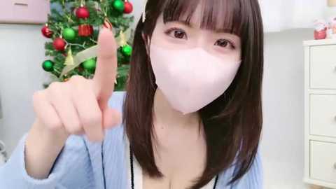 Media: Video of an Asian woman with straight brown hair, wearing a white face mask, light blue cardigan, and a white top, pointing with her left hand, in a Christmas-decorated room with a tree and white furniture.