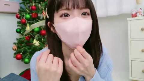 Media: Video of an Asian woman with long black hair, wearing a pink surgical mask, light blue sweater, and holding her hands near her mouth, in a room with a decorated Christmas tree and white dresser.