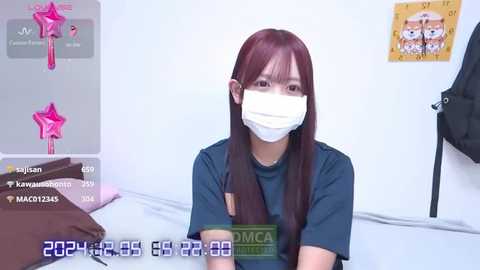 Media: Video of a young Asian woman with long, straight, dark brown hair, wearing a black t-shirt, face mask, and holding a green book. She sits on a bed with a white background, featuring pink and white decorations, and a \"Manga\" sticker.