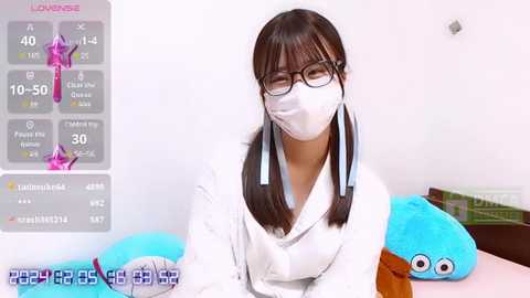 Media: A video of a young woman with long brown hair, glasses, and a white surgical mask, sitting on a bed with blue plush toys.