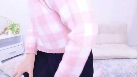 Media: A video of a person wearing a pink checkered sweater and dark skirt, sitting on a fluffy light-colored rug in a bright, minimalist room with a white couch and floral arrangement on a white dresser.