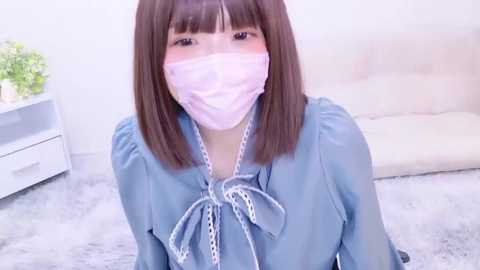 Media: Video of an Asian woman with straight brown hair and a pink face mask, wearing a light blue blouse with white lace trim, sitting on a plush gray rug in a minimalist living room.