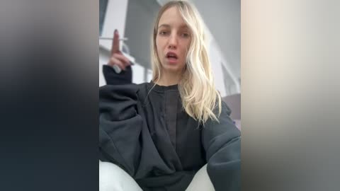 Media: Video of a blonde woman with wavy hair, wearing a black sweater, seated in a modern, minimalistic room. She gestures with her right hand, mouth open as if speaking.