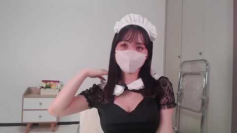 Video of an Asian woman in a black maid outfit, white headband, and mask, posing indoors with a white table and chair.