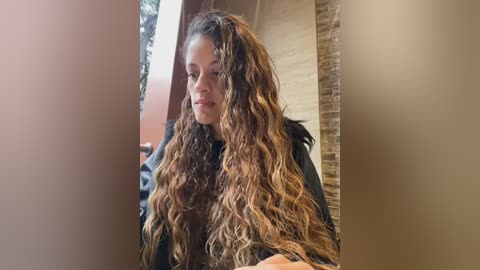 Media: Video of a young woman with long, wavy, light brown hair, wearing a black top, standing indoors near a window with a brick wall and greenery outside.