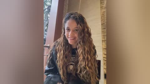 Media: Video of a smiling, light-skinned woman with wavy, long brown hair, wearing a black leather jacket with a graphic design. She is standing in a doorway, with beige walls and a wooden railing in the background.