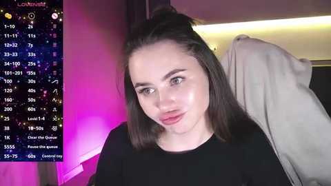 Media: Video of a young woman with straight, shoulder-length dark hair, fair skin, and light makeup, wearing a black top, indoors with a purple and pink lighting effect.