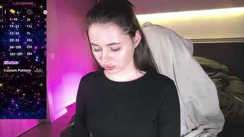 Media: Video of a young woman with fair skin and straight dark hair, wearing a black top, standing indoors. Background includes a bed with gray sheets and a nightstand with a glowing pink light.