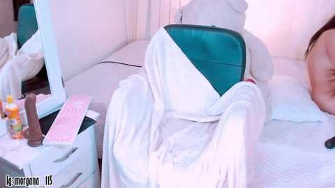 Media: A video of a woman, partially covered in white sheets, lying on a bed with a green cushion. A mirror, makeup, and toiletries are visible on a nearby white dresser.