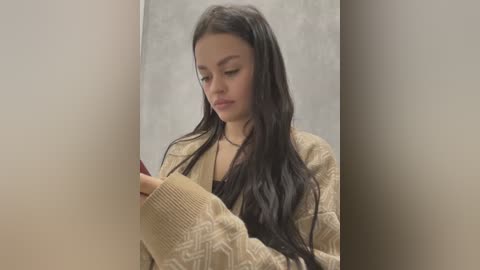 Media: Video of a young woman with long, dark hair, wearing a beige knitted cardigan and a black top, reading a book in a dimly lit room with plain walls.