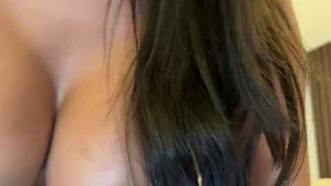 Media: Video of a close-up of a woman's bare chest with large breasts, showing dark brown, wavy hair cascading over her right shoulder. The background is blurred, revealing a beige wall and a wooden door frame.