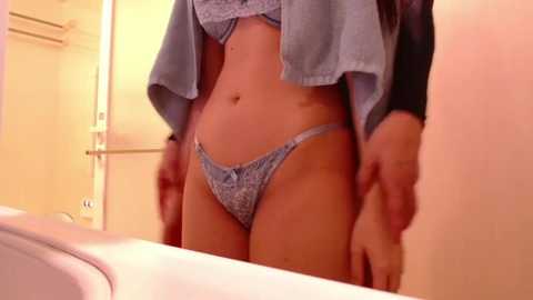 Media: Video of a light-skinned woman with long dark hair, wearing a blue lace bra and matching panties, standing in a bathroom with beige walls and a white sink.