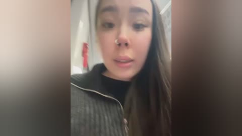 Media: A blurry video of a young woman with long brown hair, wearing a black turtleneck under a grey sweater, with a septum ring and subtle makeup, in a bathroom setting.