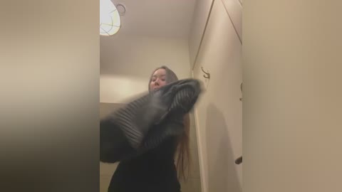 Media: Video of a woman with long brown hair, wearing a dark sweater, vigorously shaking a black dog's tail in a narrow, beige hallway.