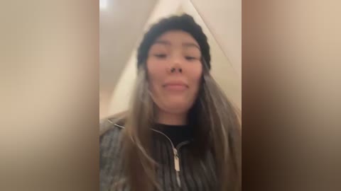 Media: Video of a young woman with light brown hair, wearing a black beanie and gray zip-up sweater, standing in a hallway with beige walls. She has a neutral expression and slightly squinted eyes.