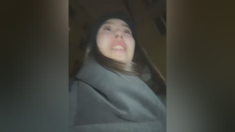 Media: A video of a young woman with fair skin, wearing a black beanie and dark coat, standing indoors with a blurred, dim background. Her expression is neutral.