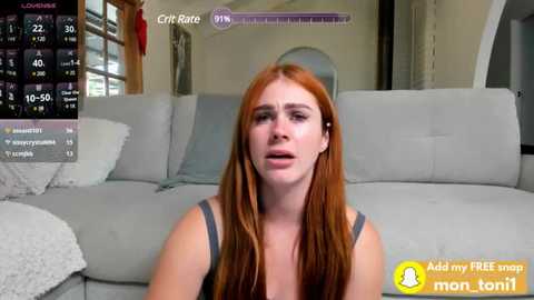 Media: Video of a fair-skinned, red-haired woman in a gray tank top, seated on a light gray sofa. The background features a modern living room with a window, a circular mirror, and a screen displaying a fitness app interface.