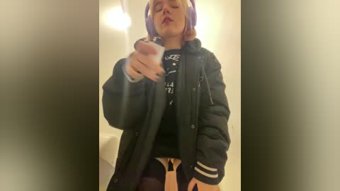 Media: Video of a woman with short, dyed purple hair, wearing a black jacket, black top, and black leggings, holding a lit cigarette, indoors with blurred background.