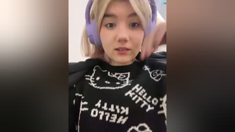 Media: Video of a young Asian woman with fair skin, wearing purple headphones, black \"HELLO KITTY\" hoodie, and a confused expression, with a hand adjusting her hair.