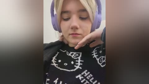 Media: A video of a young woman with light skin and blonde hair wearing large purple headphones, a black sweater with a Hello Kitty design, and a contemplative expression.