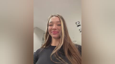 Media: Video of a young woman with long, straight, light brown hair, wearing a black top, standing in a modern, minimalist room with beige walls and recessed lighting. She has a neutral expression and slightly pursed lips.