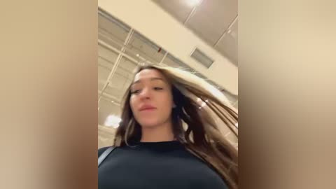 Media: A video of a young woman with long, wavy brown hair, fair skin, and a black top, taken from a low angle, with blurred ceiling lights and a neutral-toned background.