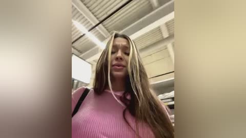 Media: Video of a young woman with long, straight, blonde hair, wearing a pink ribbed sweater. She stands in a warehouse with white ceiling panels and metal beams, with shelves and boxes in the background.