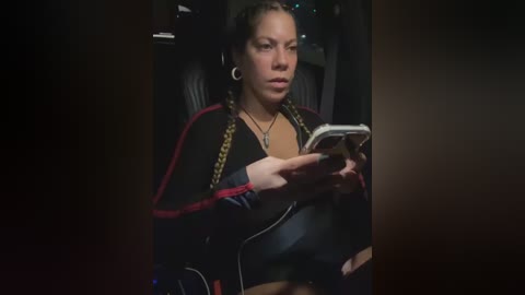 Media: Video of a woman with light skin and braided hair, wearing a black jacket with red trim, reading on a smartphone.