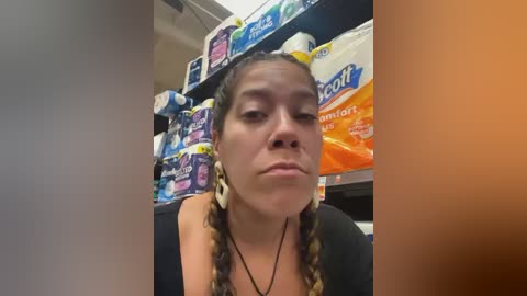 Media: Video of a Latina woman with dark braids, wearing a black top, looking serious in a grocery aisle filled with various food packages.