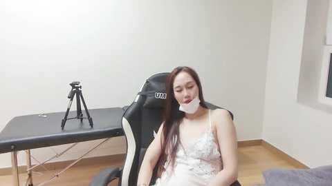 Media: Video of a young Asian woman with long, straight hair, wearing a white lace camisole, sitting on a black and white gaming chair. She has a white surgical mask on. Background includes a massage table and tripod.