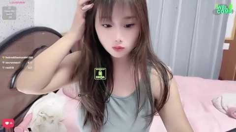 Media: Video of a young Asian woman with long, straight brown hair, wearing a grey tank top, lying on a pink bed. Background shows a wooden headboard and light grey walls.