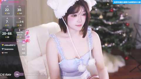 Media: A young Asian woman with fair skin and short black hair wears a light blue, frilled dress with a white cat headpiece, seated indoors next to a decorated Christmas tree.