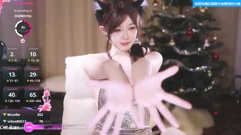 Media: A video of an Asian woman with brown hair in a ponytail, wearing a silver dress, sitting on a couch in front of a decorated Christmas tree, with a pink, translucent hand superimposed in the foreground.