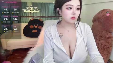 Media: A video of an Asian woman with fair skin, black hair, and large breasts, wearing a low-cut white shirt, sitting in a modern living room with plush brown furniture.