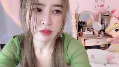 Media: Video of a young Asian woman with long, straight, light brown hair, wearing a green top. She has fair skin and red lipstick. Background shows a pink-themed bedroom with a mirror, stuffed toys, and makeup brushes.