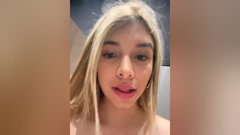 Media: Video of a young woman with light skin, straight blonde hair, and full lips. She has a neutral expression and is partially topless. Background is blurred with soft lighting.