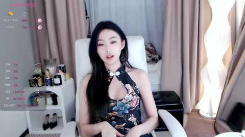 Media: A video of an East Asian woman with long black hair, fair skin, and a slender physique, wearing a floral halterneck dress, sitting in a modern living room with beige curtains and a white couch.