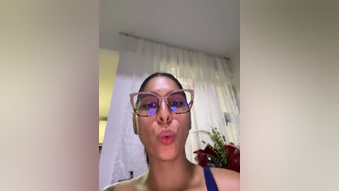 Media: A video of a light-skinned woman with straightened hair, wearing glasses and a dark blue top, making a kissy face in front of a white wall and curtains.