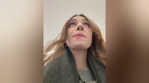 A video of a Caucasian woman with long, light brown hair, wearing a grey cardigan over a green top, looking upward, taken from a low angle.